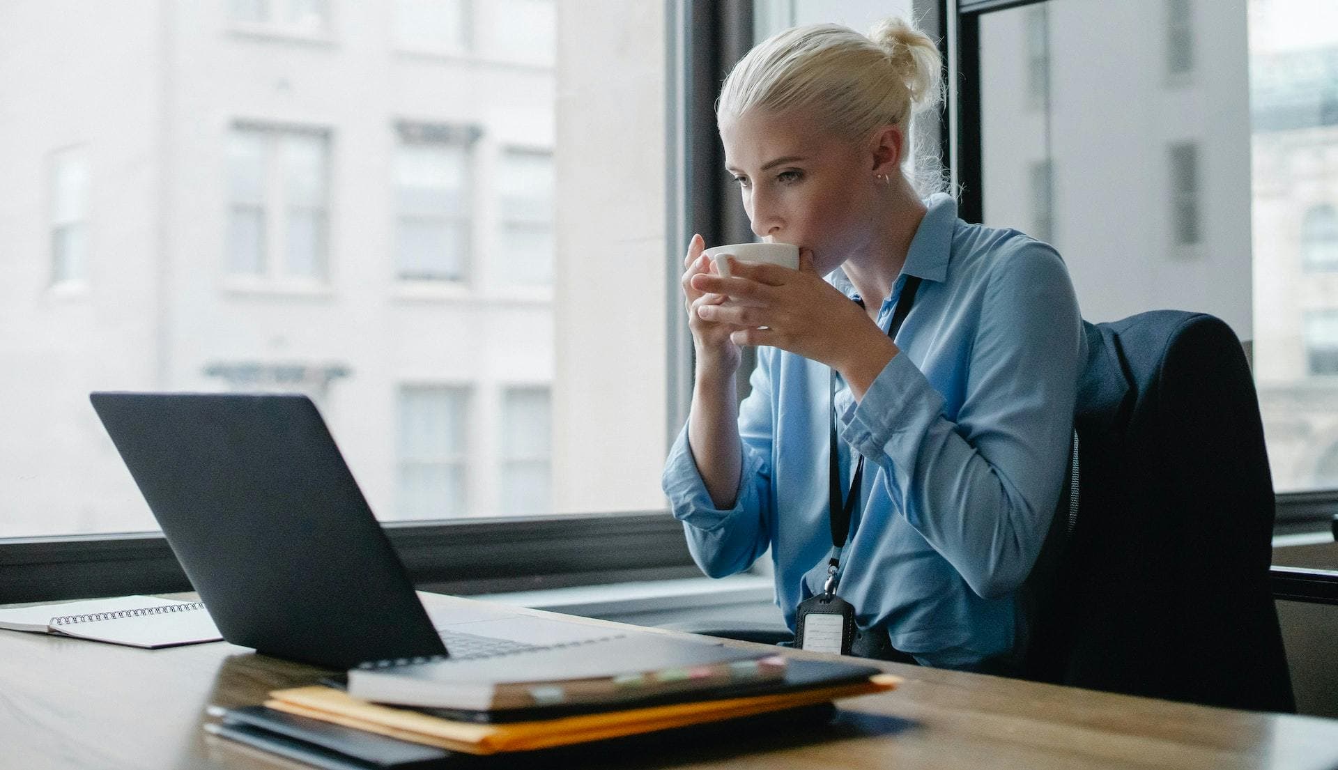 Female entrepreneur in blue shirt drinking coffee at desk in front of laptop