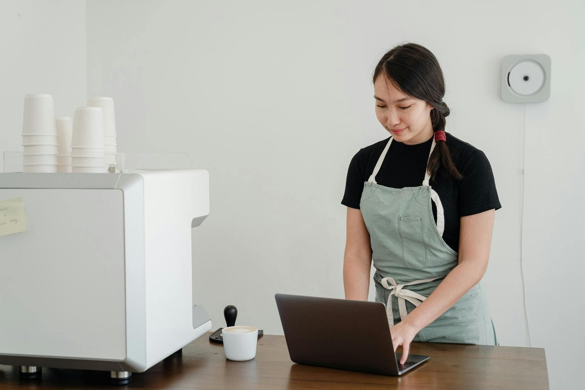 Café shop owner in apron on laptop next to coffee machine